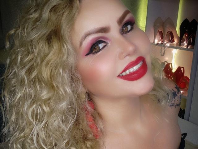 AnnaRichie with curly blonde hair, red lipstick, pink eyeshadow, and a tattoo on her right shoulder, smiling at the camera. Boobs partially visible, wearing a black top.