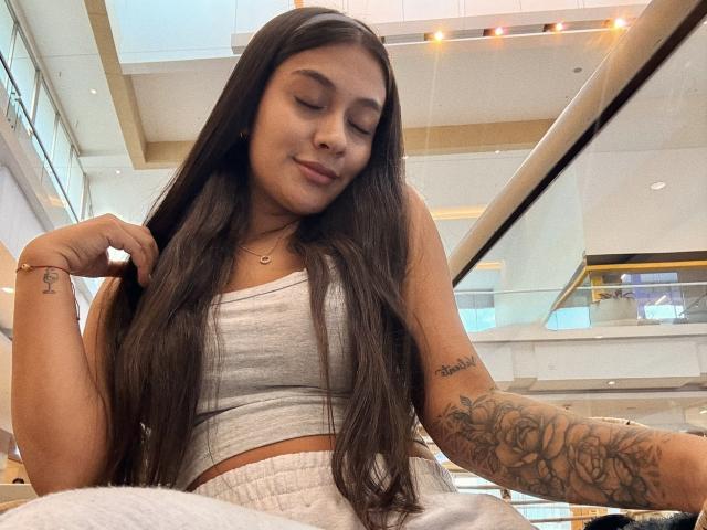 ThaliaAres has long dark hair, a cute smile, and a tattooed arm. She's wearing a gray crop top and white pants, sitting in a mall, looking down.