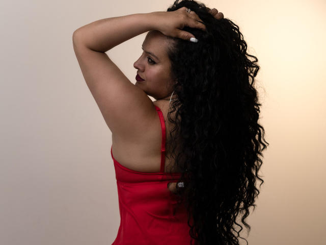 ArianaCorbyn with long curly black hair, red dress, arms raised, showing her ass, wearing dark red lipstick, against a plain beige background.