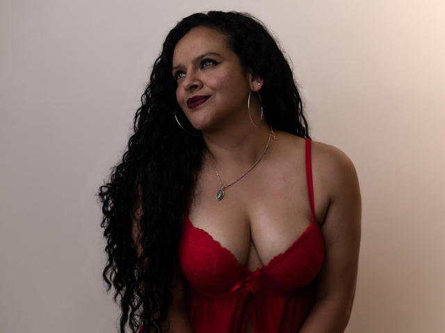 ArianaCorbyn with curly black hair, red lace bra, showing her tits, wearing hoop earrings, and a necklace, smiling at the camera. She's got dark red lipstick.