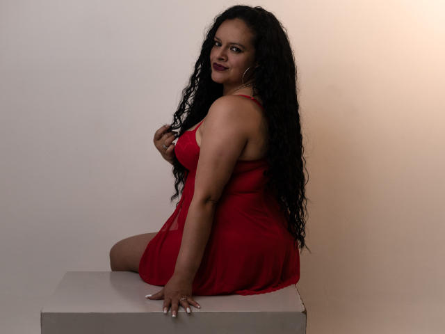ArianaCorbyn with long curly black hair, red dress, sitting on white box, looking over shoulder, smiling, dark red lipstick, hoop earrings, right hand touching hair, visible ass.