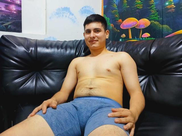 MateoSpicy, ohne Hemd, mit leichtem Bauch, in blauen Shorts, sitzt auf einem schwarzen Ledersofa, die Hände auf den Oberschenkeln, kurze schwarze Haare, kleine Brustwarzen, lächelt, helle Haut, entspannt..