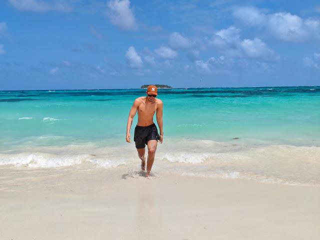 Mattew Scott, torse nu, vêtu de shorts noirs et portant une casquette orange, marchant sur une plage de sable avec une eau turquoise claire, un ciel bleu et des nuages blancs épars en arrière-plan..