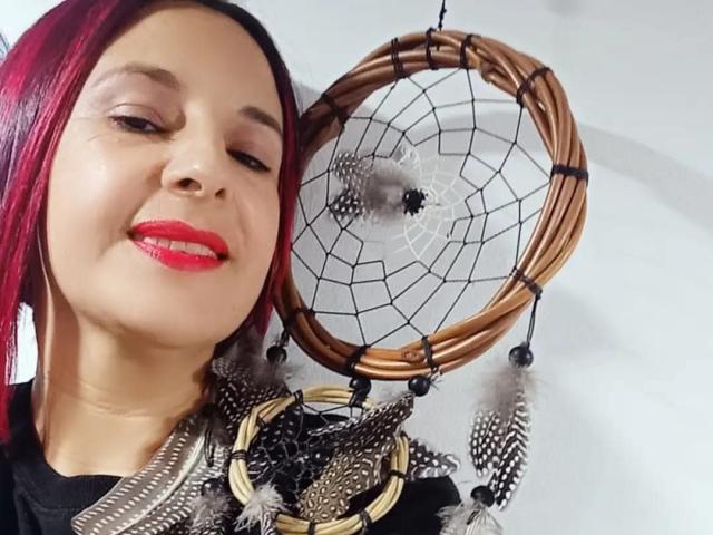 IntirRaymi, con rossetto rosso e un sorriso, è accanto a un portafortuna di legno con una rete nera e piume. Si vedono i suoi capelli neri e la sua pelle chiara. Indossa una maglietta nera..