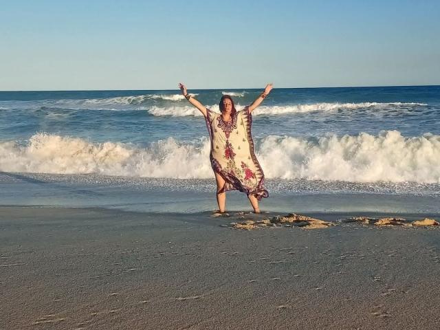 IntirRaymi se tient sur une plage de sable, les bras tendus, vêtue d'une robe fluide à motifs floraux, avec de douces vagues en arrière-plan et un ciel bleu clair au-dessus..