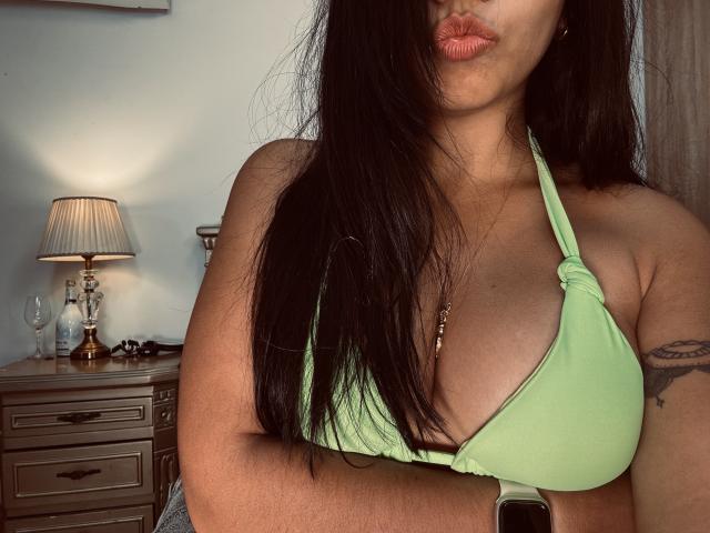 SofieWhite with long black hair, pouty pink lips, green halter top showing her big tits, tattoo on right arm, wearing a smartwatch, standing in a dimly lit room.
