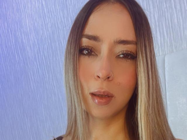 AlinaSmiith has straight, blonde-highlighted hair, brown eyes, long eyelashes, a nose ring, and glossy pink lips. Her face is centered, looking slightly to the side.
