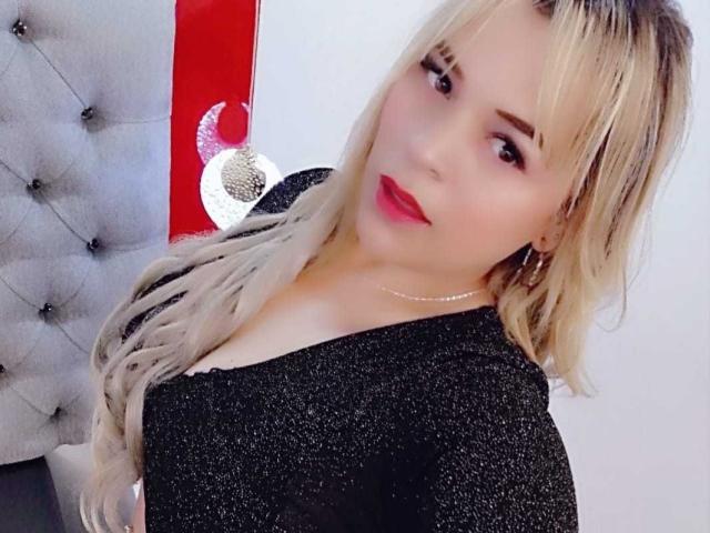 LilithRayne has blonde hair, red lipstick, and wears a black glitter top with a low neckline, showing her boobs. She's smiling slightly, with a pearl necklace.