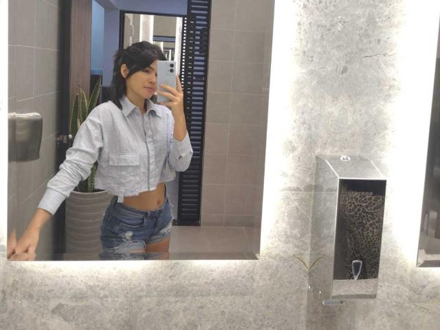 ScarlletZhen with short black hair, light gray shirt, and ripped blue denim shorts, holding a phone, showing a flat stomach and a small ass, in a bathroom mirror selfie.