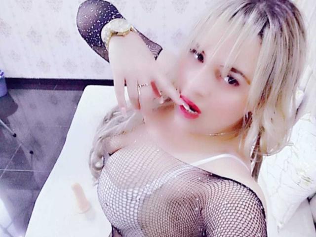 LilithRayne, con cabello rubio, top de rejilla y lápiz labial rojo, señalando su boca, mostrando sus grandes senos y un juguete sexual rosa en una cama blanca..