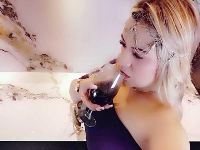LilithRayne mit blonden Haaren, die eine Kette als Kopfschmuck trägt, trinkt tiefroten Wein aus einem Glas und zeigt dabei ihre nackten Schultern und ihr violettes Kleid, während sie nach unten schaut..