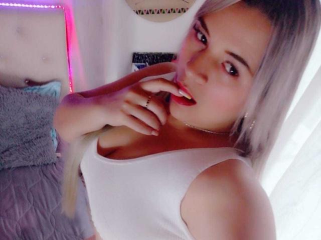 LilithRayne, con cabello rubio, camiseta blanca y lápiz labial rojo, mordiéndose el dedo, mostrando sus grandes senos, en un selfie. Su mano está cerca de su boca, luciendo linda..