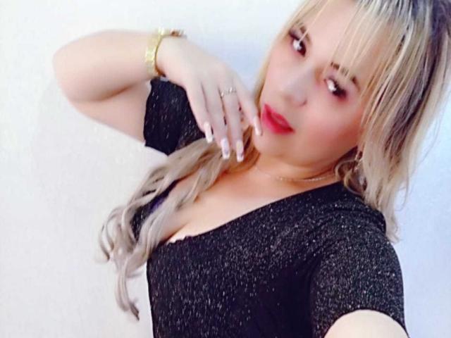 LilithRayne, con cabello rubio recogido en una coleta, vistiendo una blusa negra brillante, lápiz labial rojo y una pulsera dorada, tocándose el cuello, mostrando sus pechos..