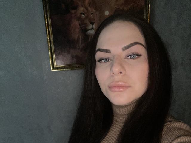 Dolci tiene la piel clara, cejas oscuras, ojos azules y labios rosados y brillantes. Lleva un cuello alto, tiene el pelo largo y negro, y un piercing en la nariz. Detrás de ella hay una pintura de un león..