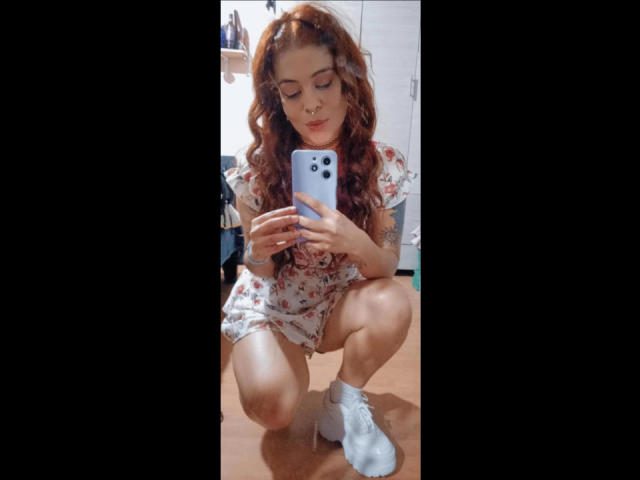 AlaiaLove, sentada no chão, com cabelo ruivo cacheado, vestido floral e meias brancas, segurando um telefone, olhando para ele, com uma tatuagem visível no braço esquerdo, em uma selfie no espelho..