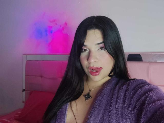 AntonelaPark with long black hair, purple fuzzy robe, pink lipstick, and necklace, pouting slightly, pink and blue neon light in background, red bed, white headboard.