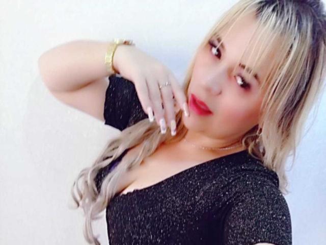 LilithRayne with blonde hair, red lipstick, black sparkly top, gold bracelet, hand on chest, slightly tilted head, soft smile, wearing ring, light skin, blonde highlights.