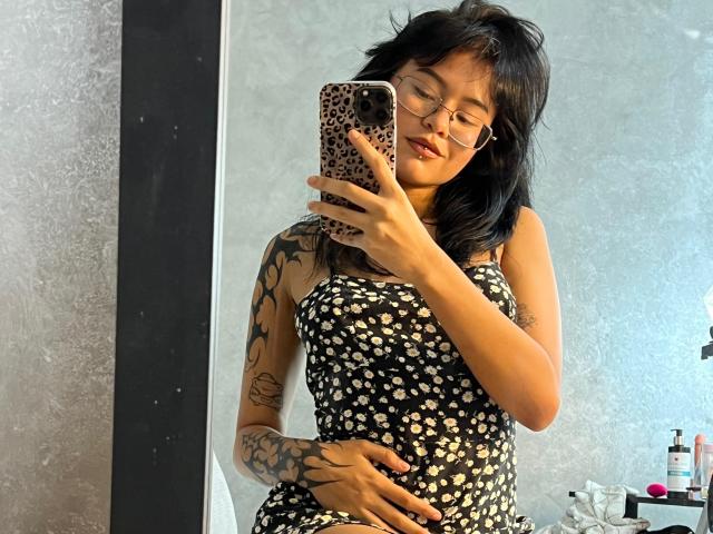 Elizabeth Roux, con gli occhiali, capelli neri, tatuaggi, indossa un abito floreale nero, tiene una cover per telefono a forma di leopardo, sta scattando un selfie allo specchio, mostrando il seno, con una mano sulla vulva..