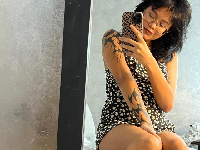 ElizabethRoux with tattoos on her arm and thigh, wearing glasses and a black polka dot dress, taking a mirror selfie with a leopard print phone case.