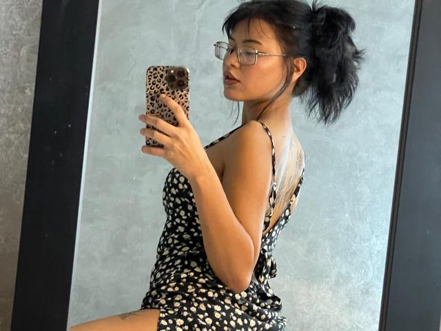 ElizabethRoux with black puffy ponytail, glasses, wearing a leopard-print dress, taking a mirror selfie, showing her back and tattoo on right thigh, holding a leopard-print phone.