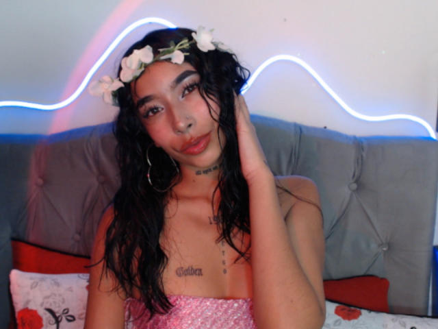 MaddyParker with wet black hair, flower crown, hoop earrings, pink sequined top, hand in hair, tattooed chest, blue neon strip, gray tufted headboard, red and white pillows.