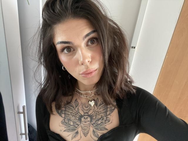 NovaSin with dark brown wavy hair, wearing a black top, showing a large tattoo of a bee on her chest, and a silver chain necklace, looking at the camera.