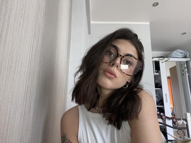 NovaSin with wavy dark brown hair, wearing round glasses, white sleeveless top, nose ring, and necklace, has a tattoo on her left arm, looking at the camera.