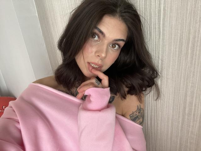 NovaSin with shoulder-length dark brown hair, pink off-shoulder sweater, tattoos on right arm, finger in mouth, brown eyes, nose piercing, soft expression, light skin, standing in front.
