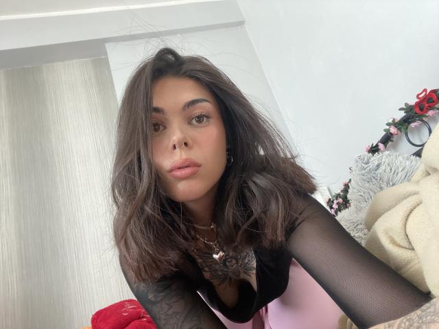 NovaSin with medium brown hair, black sheer sleeves, pink and black top, tattoos on right arm, nose ring, looking at camera, slight pout, soft light, bedroom setting.