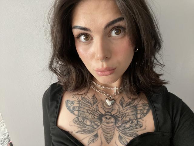 NovaSin has dark brown wavy hair, brown eyes, and a nose ring. She's wearing a black top, showing a detailed bee tattoo on her chest with heart necklace.