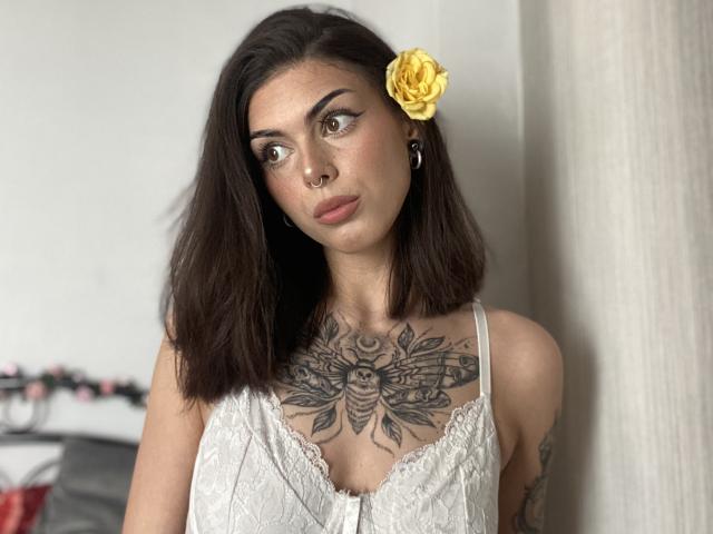 NovaSin with dark brown hair, yellow rose in her hair, nose ring, tattoos on chest, wearing white lace dress, looking to the side, standing indoors, subtle makeup, small earrings.