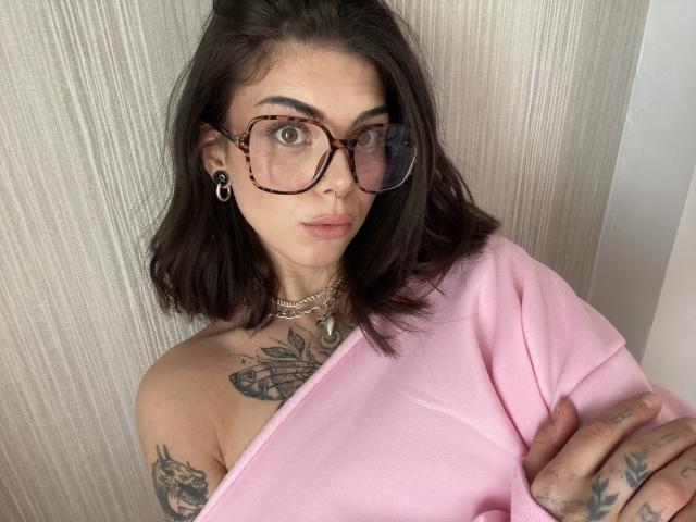 NovaSin with dark brown hair, tortoiseshell glasses, nose ring, black ear gauges, pink one-shoulder top, tattoos, chain necklace, and crossed arms, showing off her tits.