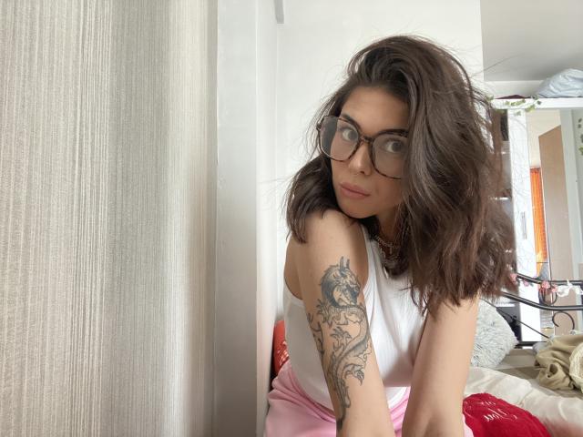NovaSin with wavy brown hair, glasses, white tank top, pink pants, tattoo on left arm, leaning on window, looking at camera, slightly smiling, natural light.