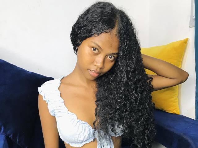 Saloma with long, curly black hair, wearing a light blue, ruffled top, has small tits, dark brown skin, and a serious expression, sitting on a blue and yellow couch.