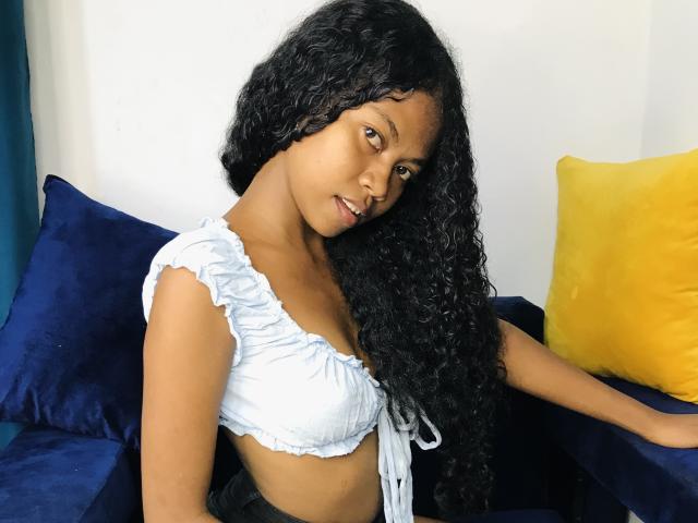 Saloma mit langen, lockigen, schwarzen Haaren, hellbrauner Haut und kleinen Brüsten, trägt ein hellblaues Crop-Top und sitzt auf einem blau-gelben Sofa. Sie schaut in die Kamera..