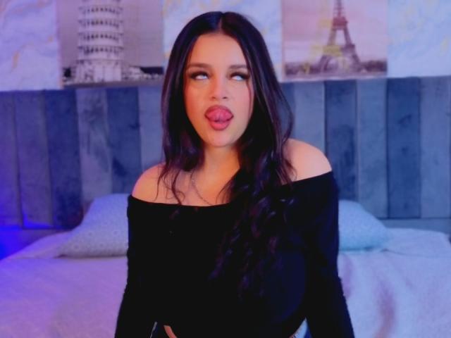 FabiValdez has long dark hair, an off-shoulder black top, and sticks her tongue out. She's looking to the side with a slightly open-mouthed, playful expression.