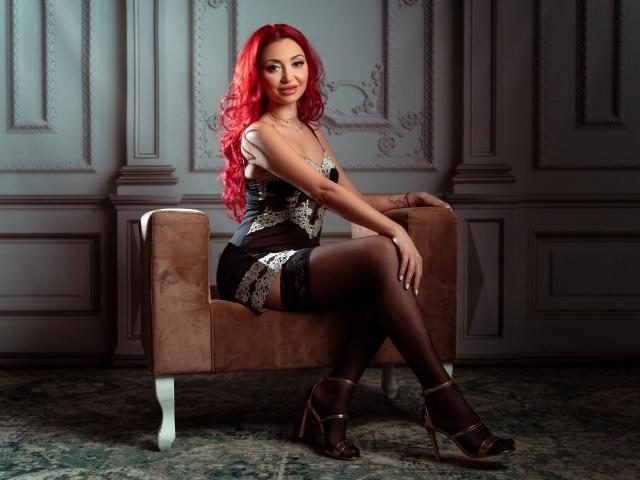 CelineHinton with red hair, black lace corset, and stockings, sitting on a brown chair, showing her ass in black high heels, looking sexy.