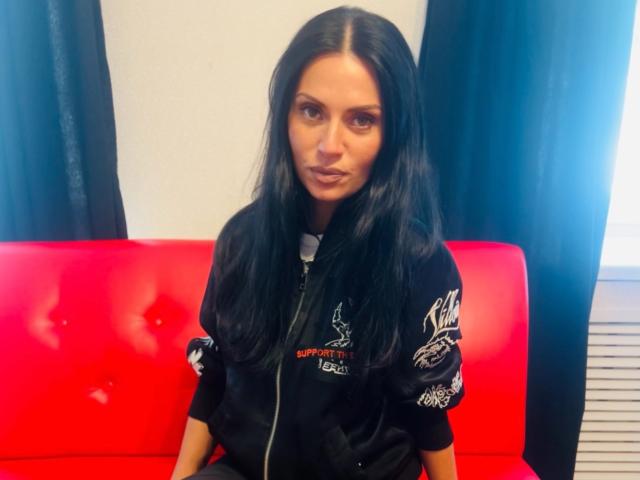 MonicaLover has long black hair, brown eyes, and a pouty face, wearing a black zip-up jacket with white and red embroidery, sitting on a red couch.
