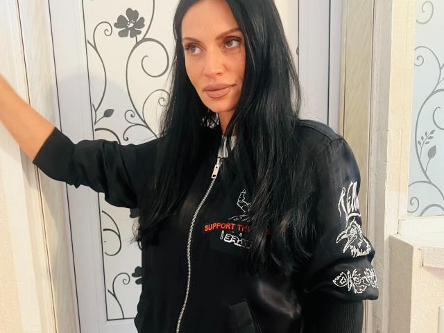 MonicaLover has long black hair, light skin, and blue eyes. She's wearing a black zip-up jacket with white embroidery, standing with one arm raised. She's looking to the side.