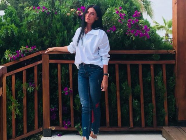 MonicaLover stands with long black hair, wearing a white button-up shirt and ripped blue jeans, leaning on a wooden railing, surrounded by purple flowers and green foliage.