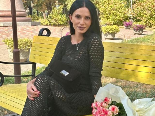 MonicaLover with black, see-through dress, black belt, and long black hair, holding pink roses, sitting on yellow bench, looking at camera, in sunny park.