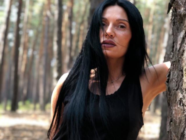 MonicaLover with long black hair, black sleeveless top, leaning against tree in forest, serious expression, visible ass, subtle makeup, light skin, slender, standing, natural light, daytime.