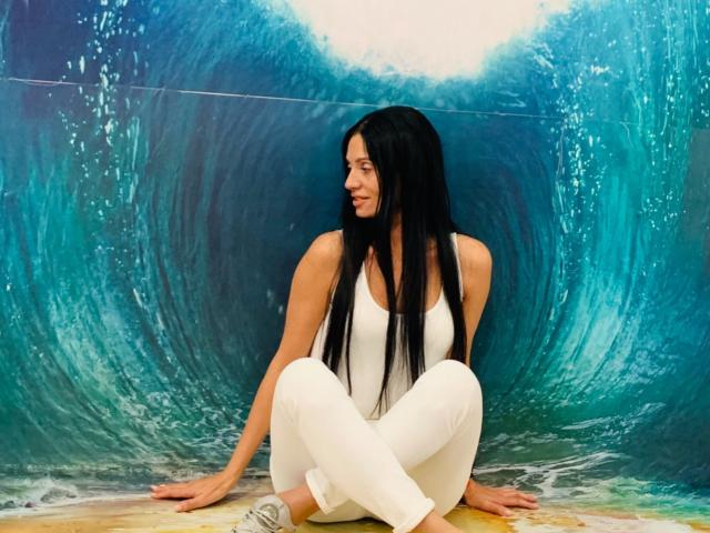MonicaLover with long black hair, white tank top, and white pants, sitting cross-legged, looking to the side, against a blue wave mural, wearing gray sneakers.