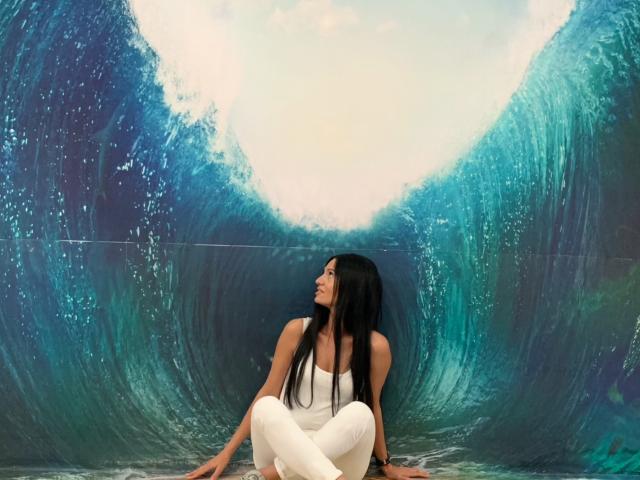 MonicaLover with long black hair, wearing a white tank top and white pants, sits cross-legged in front of a massive blue-green wave. She looks up with a relaxed expression.