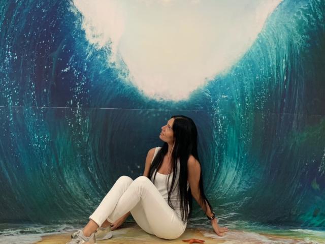 MonicaLover with long black hair, white tank top, and white pants, sitting against a giant blue wave, looking up with a relaxed, dreamy expression, wearing silver flats.