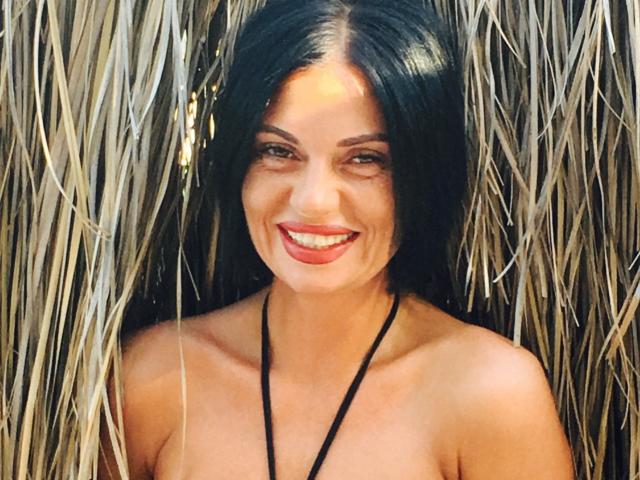 MonicaLover with long black hair, smiling with red lipstick, topless, black necklace, standing in front of straw wall. Her tits are visible, and she's looking cute.