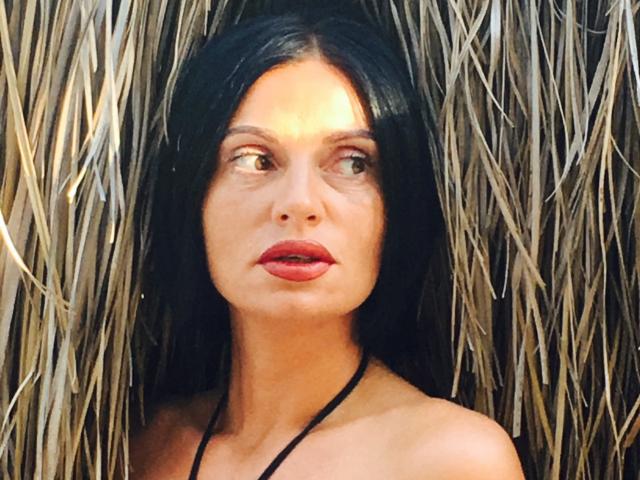 MonicaLover has dark, straight hair, red lips, and a black halter top, with her tan skin and brown eyes looking to the side, standing in front of straw.