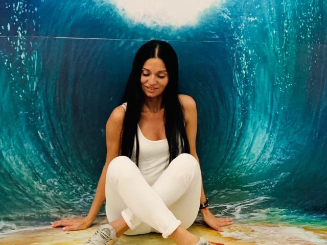 MonicaLover with long black hair, white tank top, and white pants, sitting with legs crossed, smiling, against a blue wave wall, wearing a black wristband.