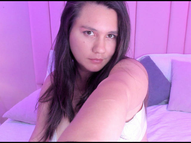 AvienLume mit langen, braunen Haaren, einem rosafarbenen Teint und einem weißen Tanktop, macht ein Selfie in einem violett beleuchteten Schlafzimmer und zeigt ein leichtes Lächeln und einen entspannten Gesichtsausdruck..