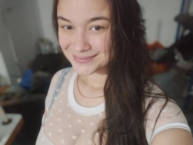 IsaLoverX, con cabello castaño largo, sonriendo, vistiendo una blusa blanca transparente con estampado de estrellas, mostrando pequeños senos, en un selfie. Luz suave, ligero rubor, expresión relajada..